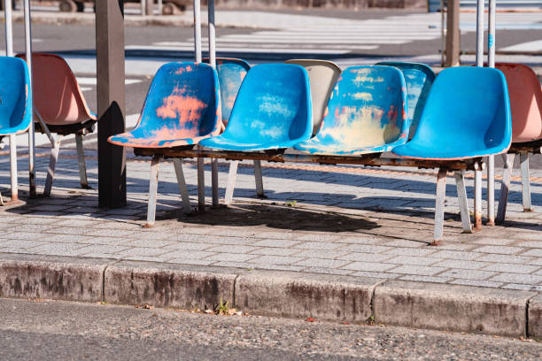 Seats at a bus stop showing signs of aging and wear, with patches of paint peeling off and fading. These plastic chairs in various shades of blue and pink sit under an overhead shelter, positioned along a paved sidewalk. The background features a blurred street view, indicating an urban setting.