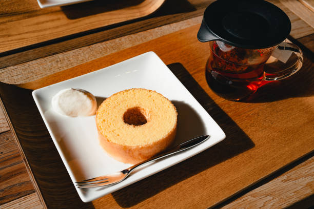 A slice of Baumkuchen cake served on a white plate accompanied by a dollop of cream and a fork, placed on a wooden tray. A pot of freshly brewed tea in a glass pot with a black lid is also on the tray, creating a cozy and inviting scene.