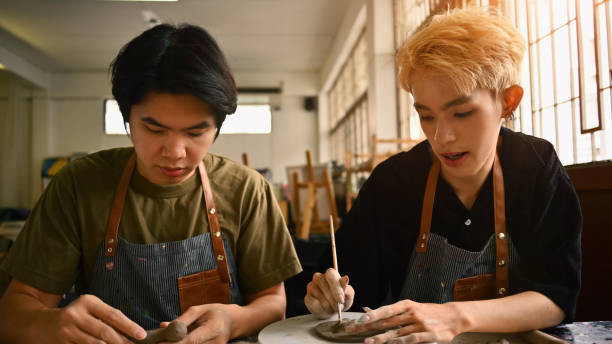 istock Two art students working on pottery projects in a studio. Both are focused, wearing aprons, and shaping clay with their hands and tools. The workspace is filled with natural light and art supplies 2158718193