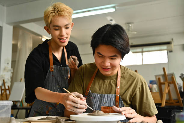 istock Two art students, one sculpting clay on a pottery wheel while the other smiles and offers help, surrounded by art tools and supplies in a creative studio, Crafts and DIY with people concept 2158718726
