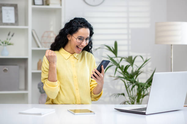 istock Excited woman celebrating success while using smartphone and laptop in modern office 2158721895