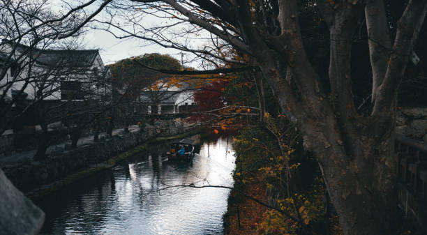 Peaceful moment captured of a leisurely boat ride on a narrow canal surrounded by autumn foliage. The water reflects the vibrant colors of the season, while the bare branches of nearby trees frame the scene. Traditional architecture lines the canal, adding to the serene atmosphere.
