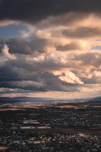 Elevated view of a city under a dramatic sky with dark rolling clouds and patches of sunlight. Surrounding mountains and plains create a striking contrast between urban and natural elements, evoking a sense of dynamic weather and expansive environment.