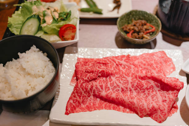 High-quality Japanese Wagyu beef slices displayed on a white plate, accompanied by a bowl of steamed white rice, fresh salad with cucumber and tomato, and a small dish of sautéed vegetables. The meal showcases traditional Japanese cuisine, focusing on premium beef and fresh ingredients.