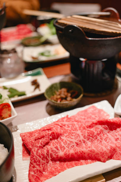 High-quality marbled beef slices beautifully presented on a white plate, ready for a traditional Japanese hot pot meal. The setting features a pot and various dishes in the background.