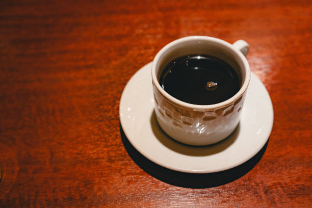 A close-up view of a white cup filled with black coffee, placed on a matching white saucer. The warm, rich wooden tabletop provides a cozy background. The light reflects gently off the surface of the coffee.