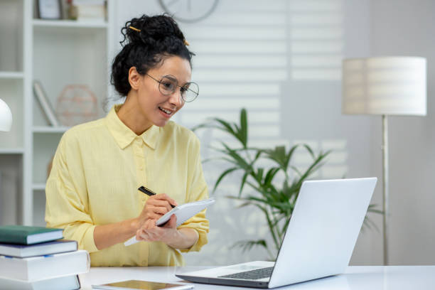 istock Woman working from home taking notes while looking at laptop in well-lit home office 2158728654