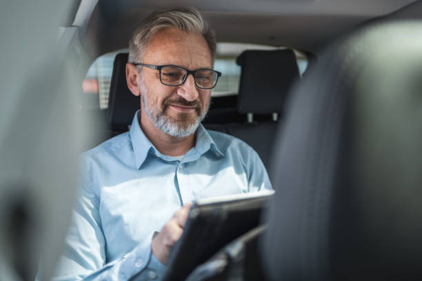 istock Mature man with glasses and beard working remotely from a taxi using a tablet. Concept of modern business commuter, remote work, and technology. Professional and relaxed atmosphere. 2158728959