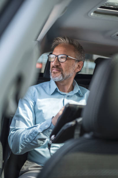 istock Mature business professional working remotely while commuting in a taxi. The image captures a man with glasses and a beard, looking out the window thoughtfully, holding a tablet and enjoying a bright day. 2158728961
