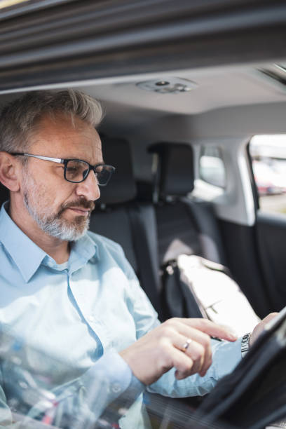 istock A mature man working remotely from a car, using a tablet. He is wearing glasses and a blue shirt, focusing on his work. This image symbolizes remote work, business commuting, and modern technologies. 2158728965