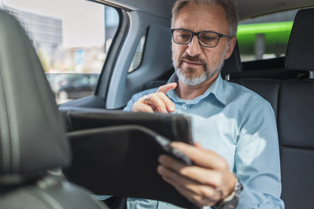istock Mature businessman working remotely on his tablet while commuting by car. He is engaged in his work, leveraging the time effectively while traveling. The urban setting in the background suggests a busy day. 2158728969