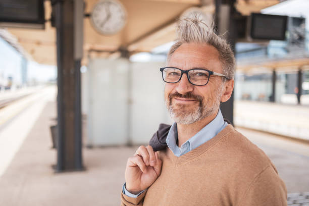 Senior man with glasses awaiting train at station, carrying jacket over shoulder. The image captures a confident, relaxed commuter during his early morning journey, signifying resilience and daily routine.