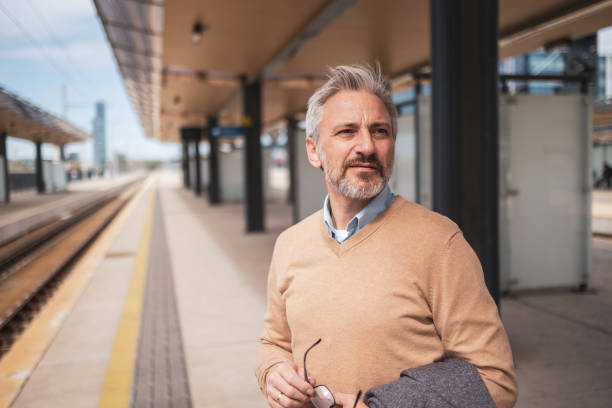 istock Mature professional man holding a coat and glasses, standing on a train station platform, looking thoughtful. He is dressed in business attire, preparing for a commute or business trip. 2158728993