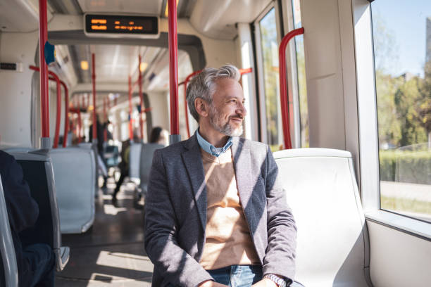 A mature man with a grey beard and a blazer smiling confidently while commuting on a train. The warm sunlight filters through the windows, creating a relaxed and content ambiance.