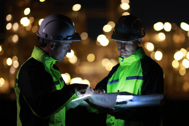 A smart chemical engineer using a digital tablet working late at night with a refinery plant in the background.Engineer working on outdoor building site at night. Engineers working late in power plant