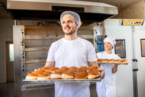 istock Male baker holding tray of bread in bakery aisle. 2158773059