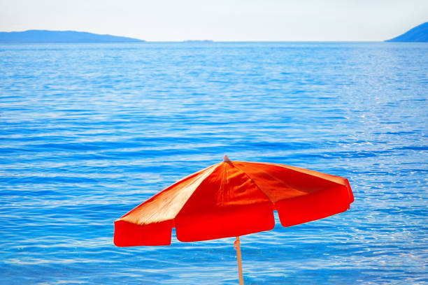 Umbrella providing shade on a serene beach and sparkling blue waves. Red umbrella is sitting on the beach next to the ocean