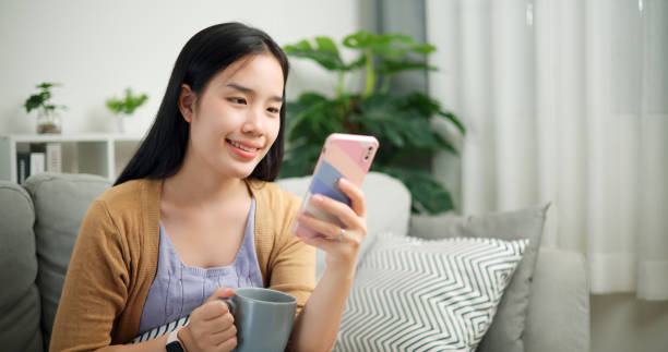 istock Relaxed woman using her mobile phone while drinking a cup of coffee on sofa at home. 2158802833