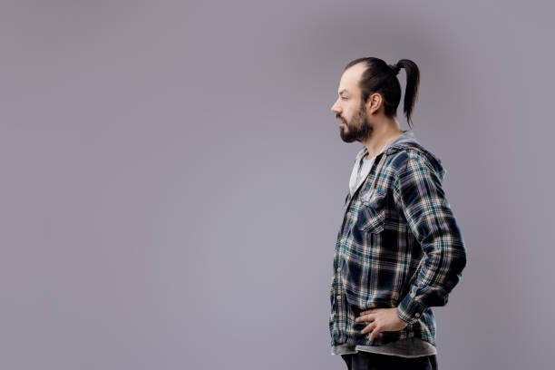 Bearded man in a plaid shirt, standing against a gray background, lost in deep thought, exuding masculinity and solitude with a trendy hipster style
