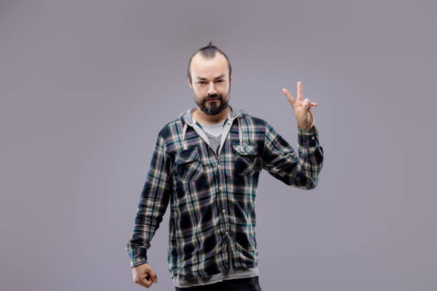 Bearded man in a plaid shirt energetically gesturing rock and roll against a gray background, exuding cool confidence and rebellion as he gazes at the camera