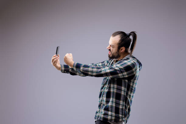 Bearded man with a ponytail shows intense emotions while using a smartphone, reflecting stress and overwhelm from technology's impact on mental health in younger generations