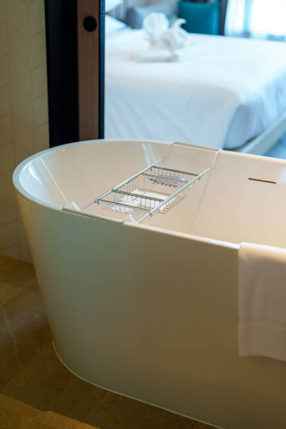 A simple, minimalist bathroom featuring a white clawfoot tub, white tile floor, white walls, and white curtains in front of a window.
