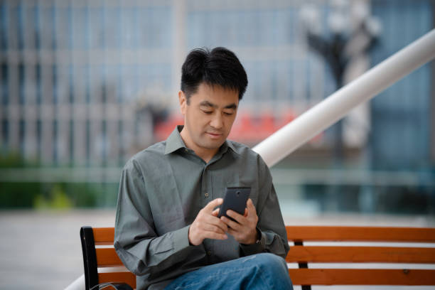 istock Middle-aged man using smartphone while sitting on bench in city street 2158827801