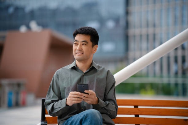 istock Middle-aged man using smartphone while sitting on bench in city street 2158827841