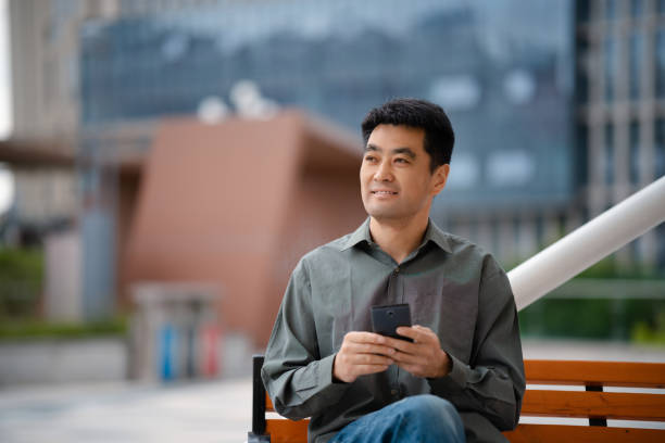 istock Middle-aged man using smartphone while sitting on bench in city street 2158827843