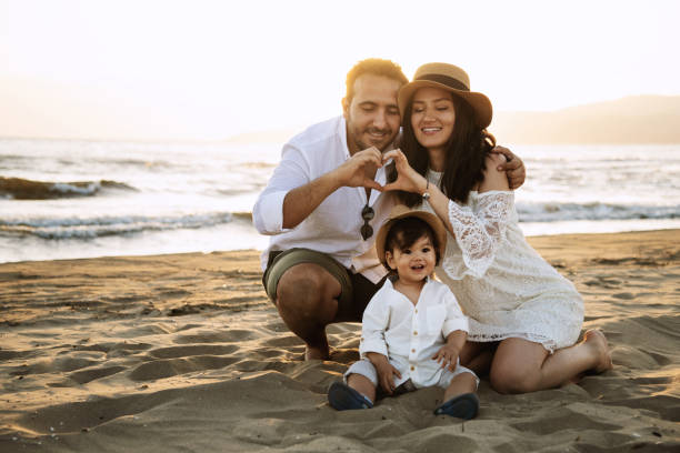 A Turkish father, Uzbek mother, and their baby boy sit on the sandy beach at sunset. All dressed in white, with the mother and baby wearing hats. They look very happy together