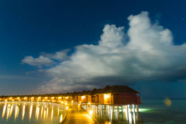 Tranquil scene of a wooden pier reaching a gazebo in the ocean, illuminated by warm lights, with palm trees framing a beautiful sunset in the Maldives