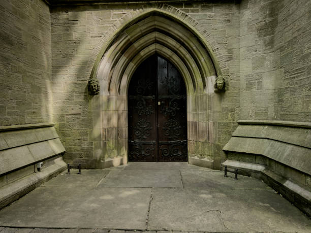 A photograph of the front entrance of St Marks Church in Worsley, Salford, Greater Manchester, England. The photograph was created on a beautiful summer day and shows two ornate, worn gargoyles on either side of the reinforced wooden doorway