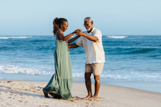 Mature African American couple dancing joyfully on beach An older African American couple enjoys a joyful moment dancing together on a sandy beach, with ocean waves in the background, emitting happiness and togetherness. black couple sunset stock pictures, royalty-free photos & images