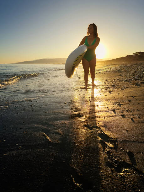 A woman in a green swimsuit stands on a sandy beach holding a paddleboard as the sun sets behind her. The golden light casts long shadows and illuminates the wet sand, creating a magical and serene atmosphere. The calm ocean waves gently lap at the shore, enhancing the peacefulness of the scene. The woman gazes thoughtfully into the distance, embodying a moment of reflection and tranquility.