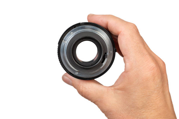 A hand holds a camera lens against a white background. The lens is black with a silver ring.