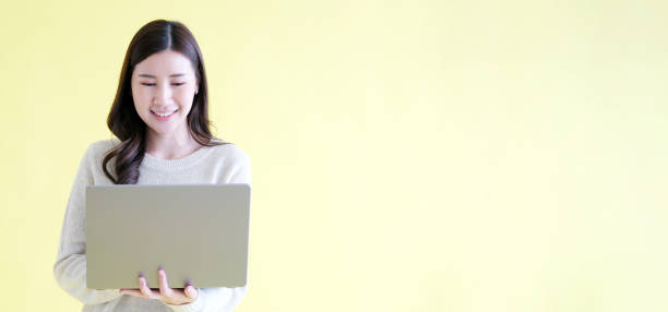 istock Young asian woman holding laptop computer, smiling and looking at camera while standing over isolated yellow background with copy space 2158947682