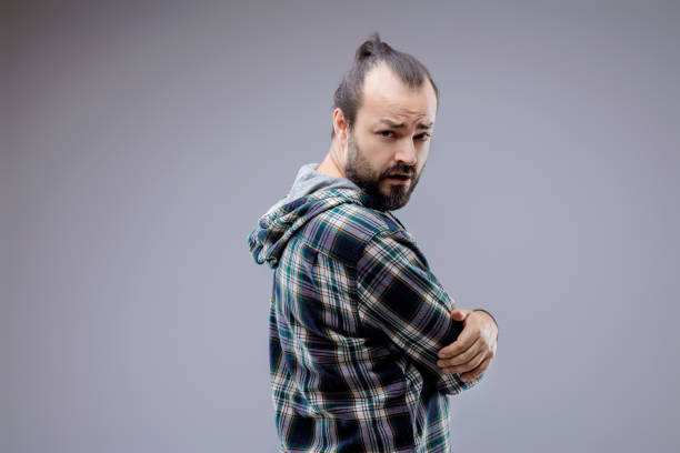 Skeptical man in his 30s with a bun and beard looks over his shoulder with crossed arms, showing suspicion and doubt in a plaid shirt and hoodie against a gray background
