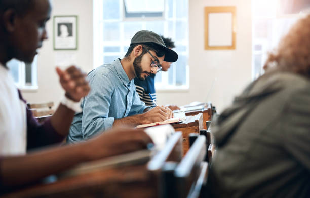 istock Classroom, university and man student with exam for education, scholarship or studying. College, knowledge and male person writing notes for learning in lecture for test or assignment at campus. 2158958400