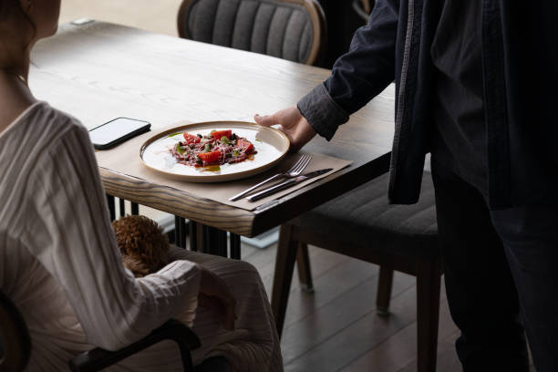 Woman sitting in modern restaurant. Waiter serving plated dish of fresh tomatoes, greens, and meat. Small dog on her lap. Eating out