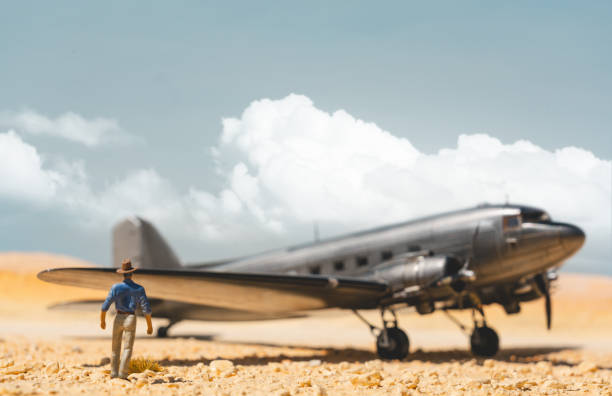 A Fedora-wearing figure approaches a scale model of the iconic bare-metal DC-3 Dakota, sitting in the desert. Scale model photography.