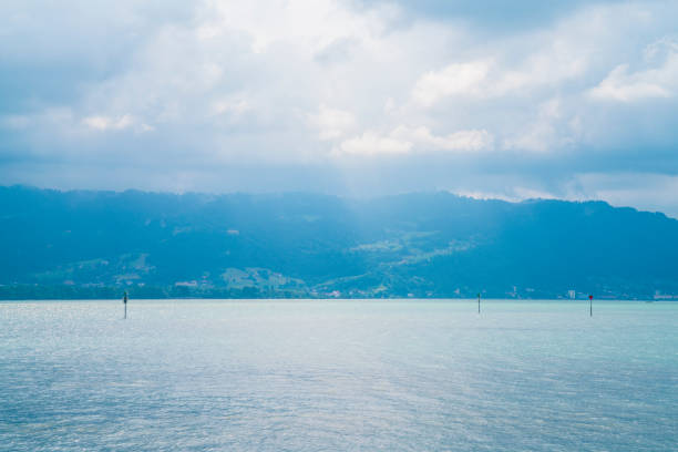 Germany, Lindau bodensee turquoise lake water view to austrian coastline, bregenz and pfaender mountain with sun and dramatic rainy thundery sky panorama landscape