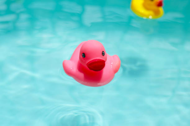 Yellow rubber duck toy sitting on a white balcony wall, facing forward with a blurred bright blue sky, the sea, and green trees in the background, creating a relaxed summer holiday mood