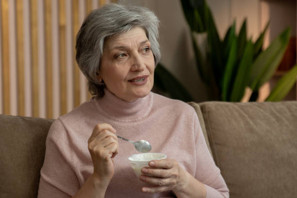 istock Elderly Woman Eats Yogurt While Sitting On A Couch In The Evening 2159003631