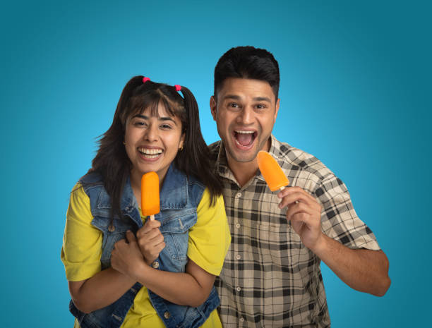 Portrait of excited young couple opening mouth and eating orange popsicles on isolated blue background