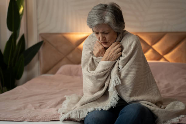A woman in her late 60s or early 70s is sitting on a bed, wrapped in a blanket, and looking unwell. She appears to be cold and may be feeling sick. stock photo