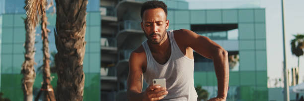 istock Young bearded male athlete is resting after training, sitting on bench, using smartphone, panorama 2159006156