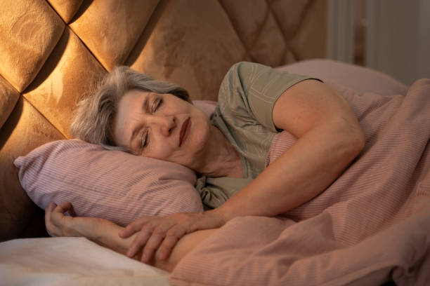 At night, an elderly woman can be seen sleeping soundly in bed. She is covered with a blanket, and her arms are wrapped around a pillow. The bed is made with pink sheets and a pink blanket. stock photo