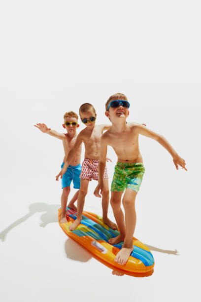 istock Three boys in swim trunks and sunglasses balancing, as surfing on inflatable board against white studio background. 2159012080