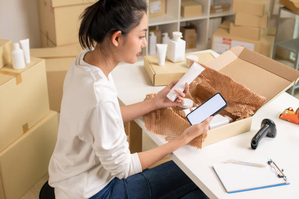 istock Asian e-commerce seller inspecting and sorting products for customers, using mock-up app, in a warehouse setting for an online beauty shop. 2159022121