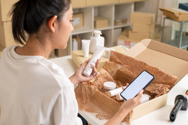 istock Asian e-commerce seller inspecting and sorting products for customers, using mock-up app, in a warehouse setting for an online beauty shop. 2159022302